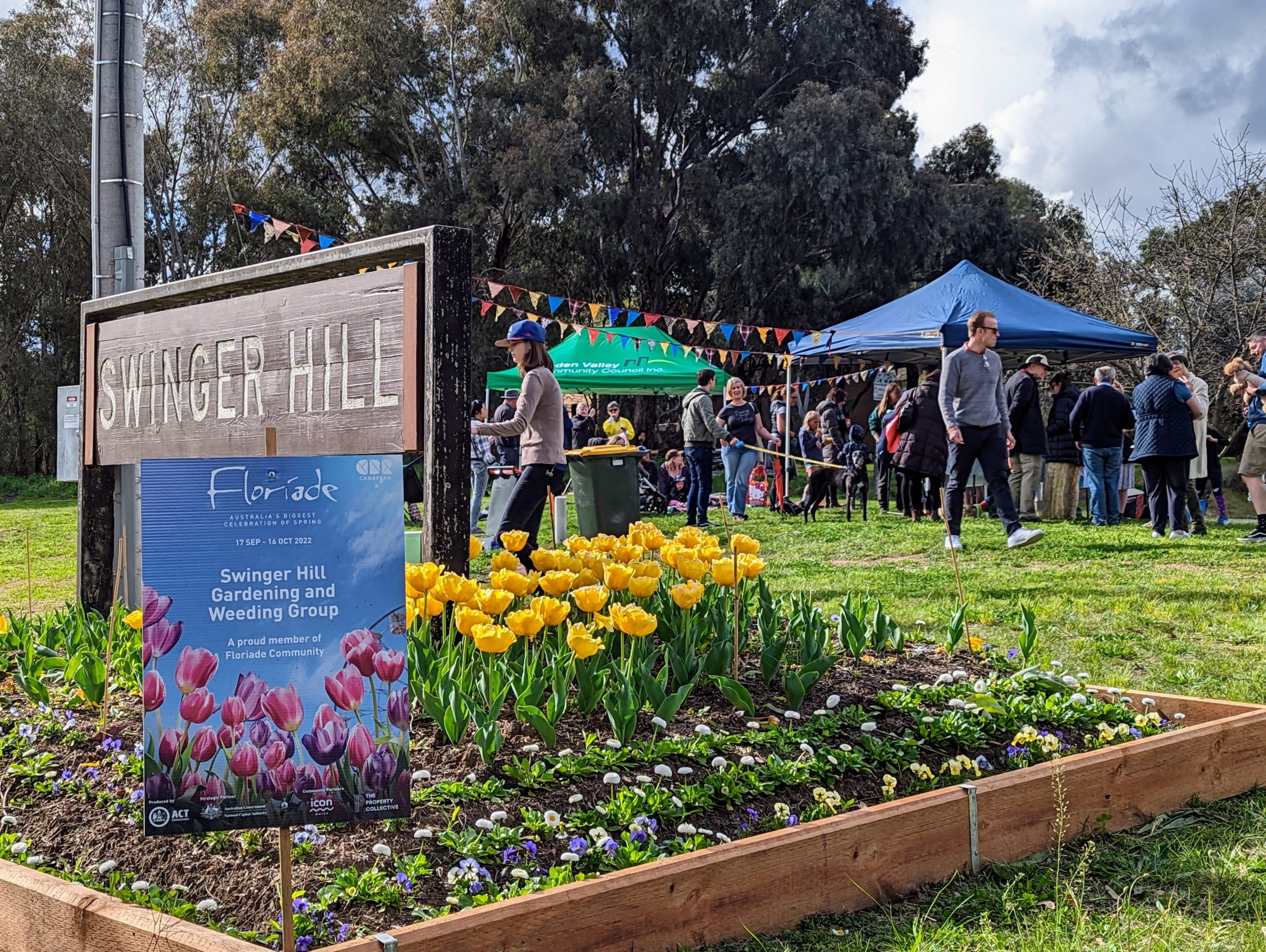 Flower bed with bright yellow tulips and mixed spring plants in the foreground, beside a wooden sign reading “Swinger Hill” and a Floriade information board, with a crowd of visitors gathered under tents and bunting in a park setting behind.
