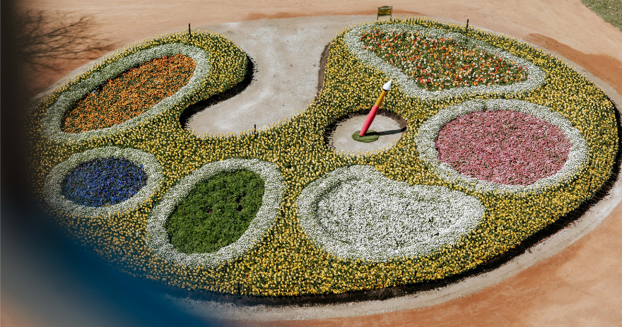 Aerial view of a large, artist’s palette‑shaped garden bed filled with patterned sections of yellow, white, pink, orange and blue flowers, with a paintbrush sculpture placed in the centre.