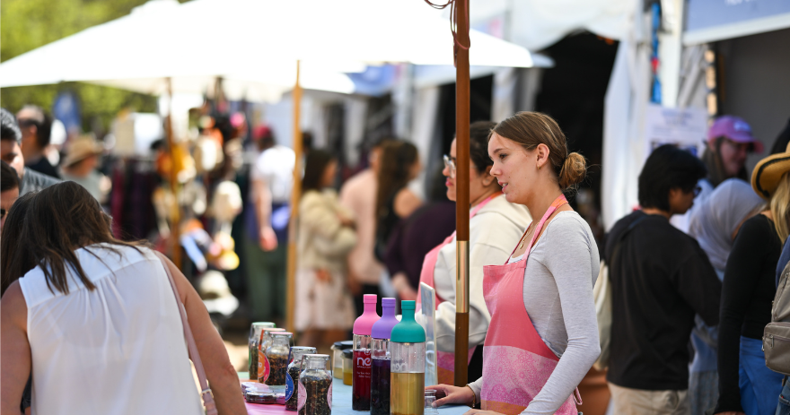 Outdoor market scene with a stallholder serving visitors beneath white tents and a busy crowd.