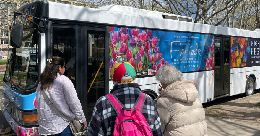 People board a bus decorated with large floral graphics parked along a street lined with trees.
