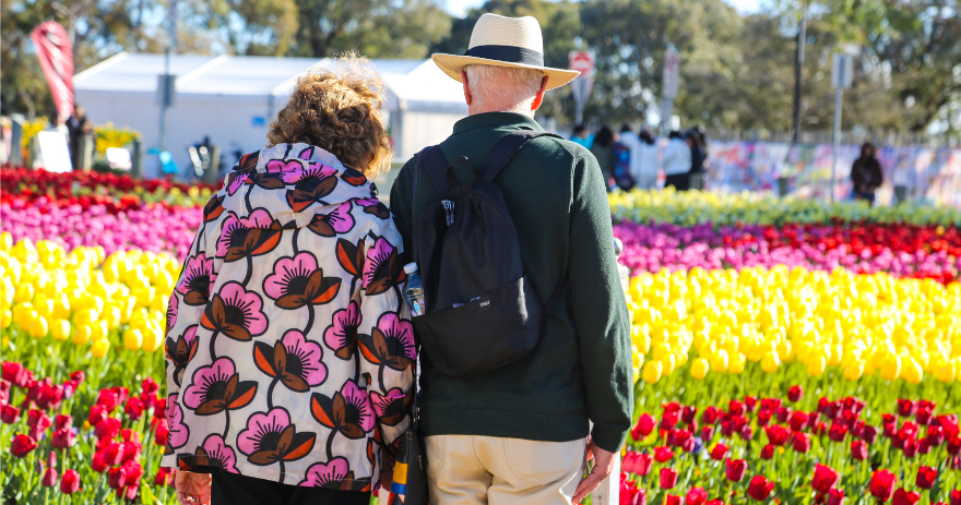 Two visitors stand side by side facing a large display of colourful tulip beds, with rows of red, yellow and pink flowers stretching across a garden.