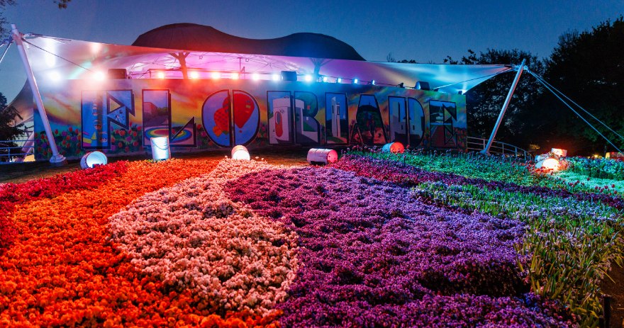 Colourful flower beds arranged in patterned rows in front of a large “FLOREADE” sign, lit with multicoloured lights at night.