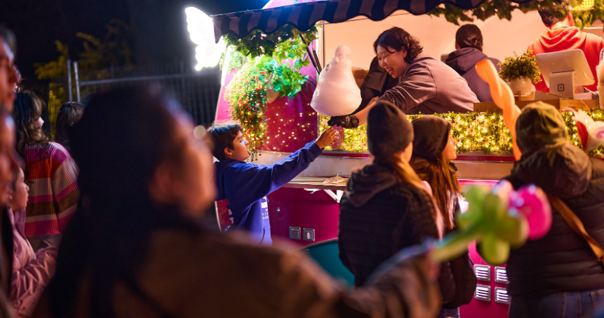 Child reaching up to receive a large piece of fairy floss from a vendor at a brightly lit food stall at night.