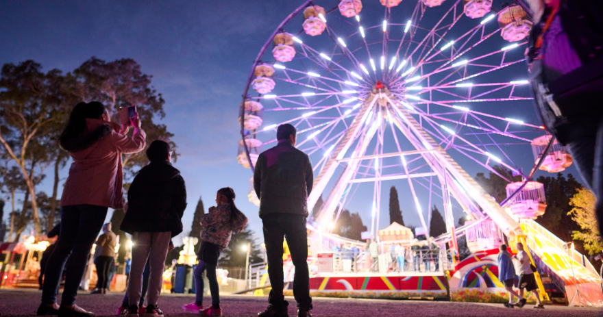 Visitors stand looking up at a large illuminated ferris wheel, with ride structures and lights visible at night.
