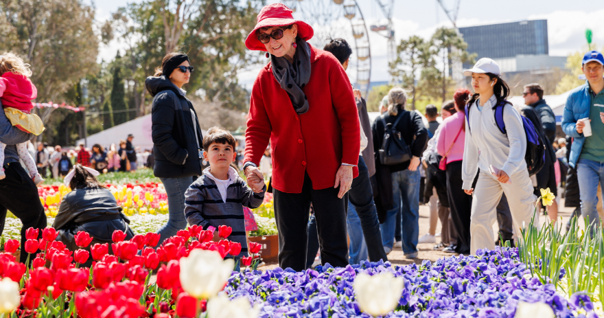 Visitor in a red hat walking through colourful flower beds while holding a child’s hand, with crowds and a ferris wheel in the background.