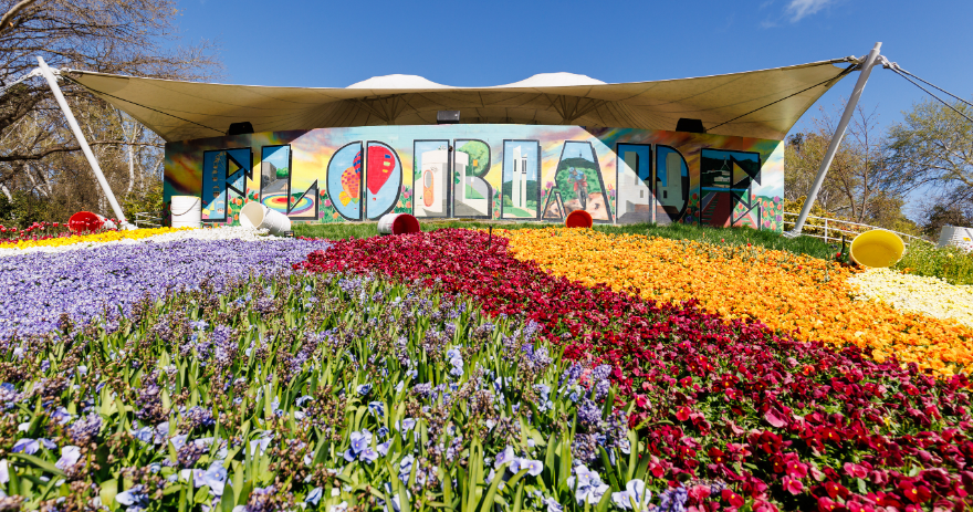 “Floriade” sign displayed across a building with large, patterned flower beds in purple, red, orange and yellow in the foreground.