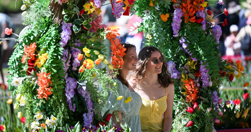 Two visitors standing beneath a decorative arch covered in greenery and bright flowers, surrounded by garden displays.