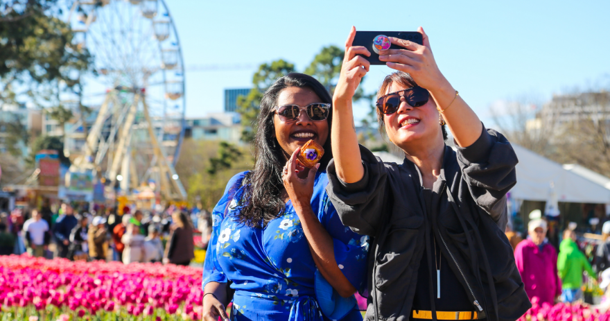 Two visitors taking a selfie in front of pink tulips, holding pastries, with a ferris wheel and crowds visible behind.