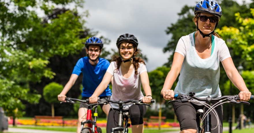 Three cyclists riding along a park path with garden beds and trees in the background under a cloudy sky.