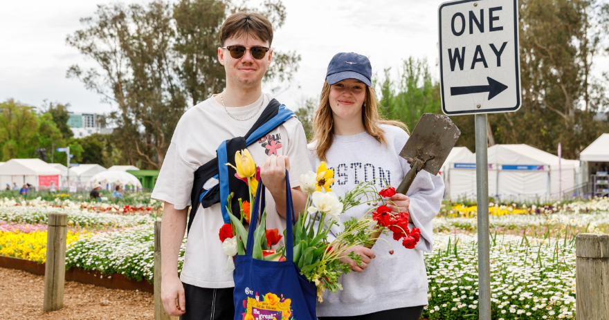 Two visitors standing in front of flower beds holding freshly picked flowers, bags filled with plants and a garden trowel visible.