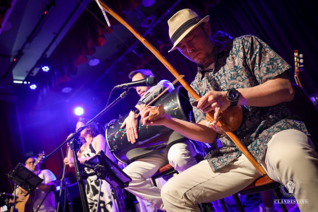 Musicians perform on stage under purple lighting, featuring a string instrument player in the foreground and percussion and vocal performers in the background.