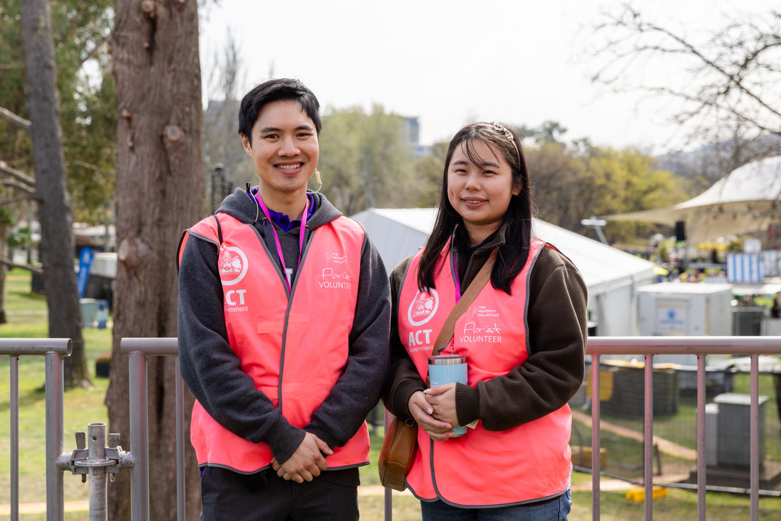 Two Floriade volunteers wearing bright pink vests stand side by side outdoors near a railing, with festival tents, trees, and garden displays visible in the background.