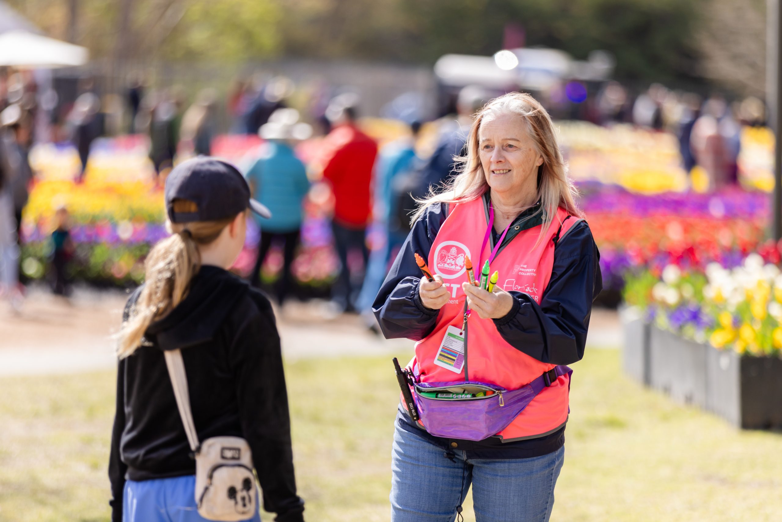 Floriade volunteer wearing a pink vest hands an information card to a child in a park filled with colourful flower beds, with visitors walking and displays in the background.