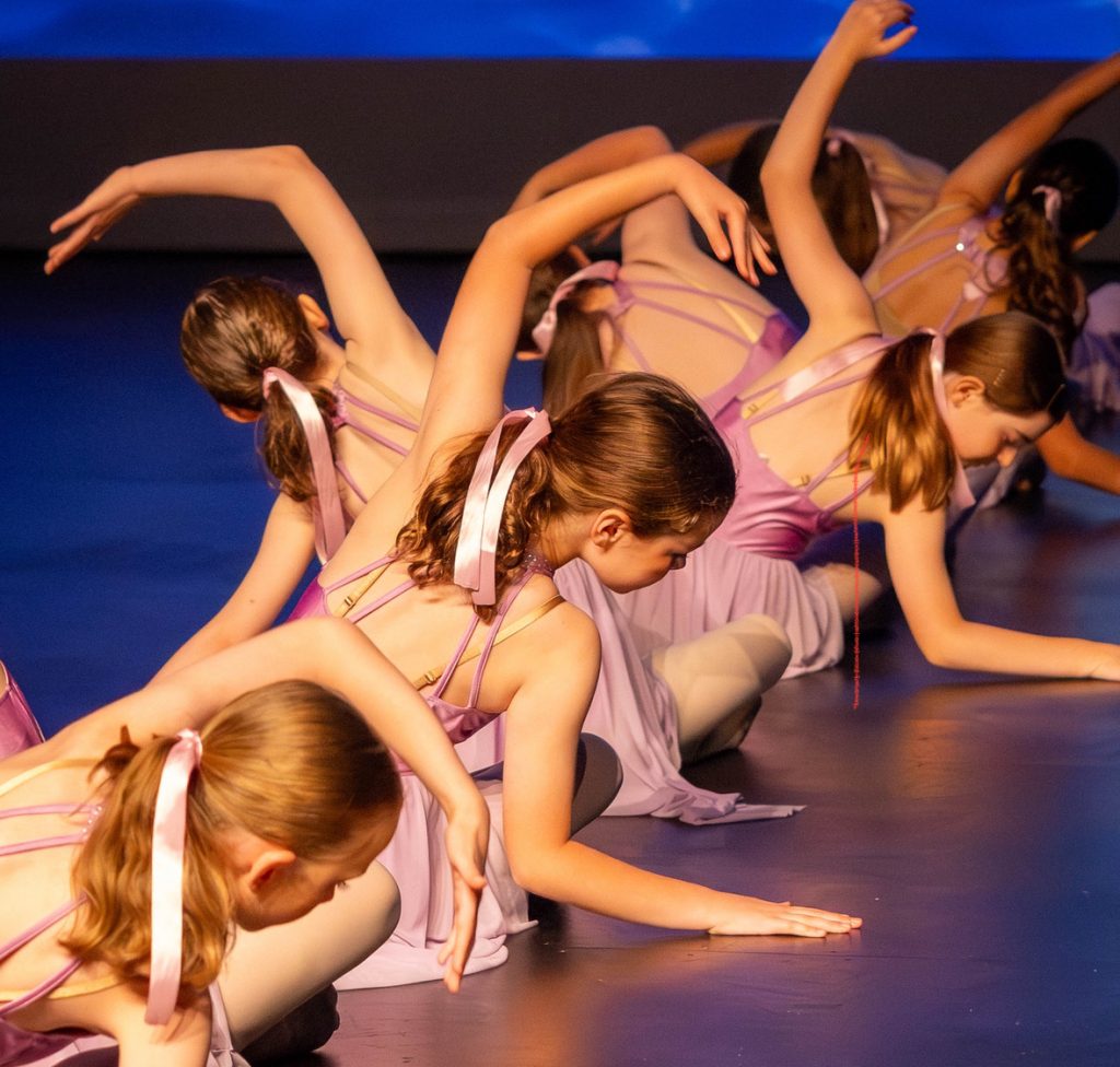 Children participating in a dance class in a mirrored studio, raising their arms during a group routine.