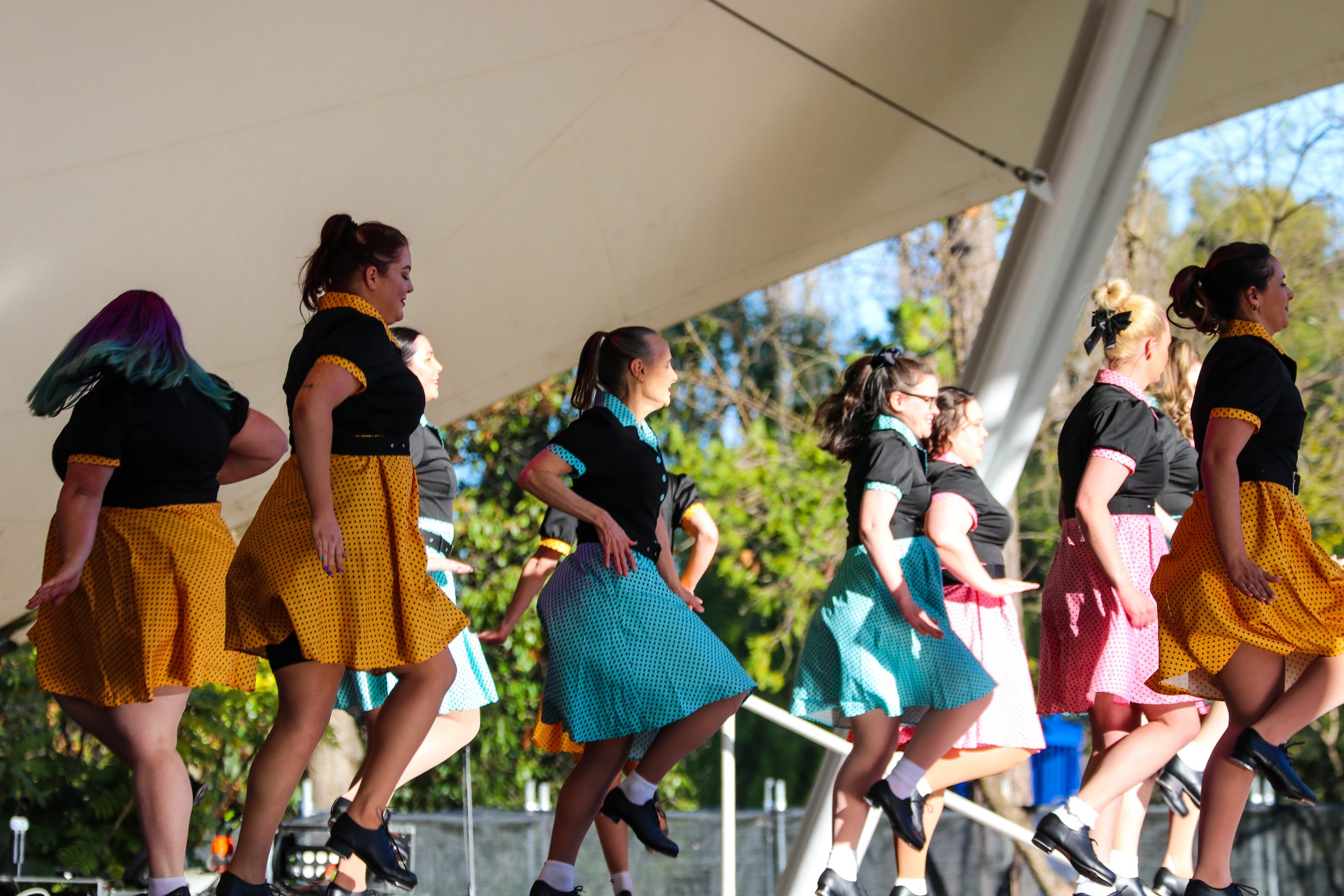 Dancers performing on an outdoor stage at Floriade, wearing coordinated outfits with bright skirts and black tops, mid‑step in a choreographed routine beneath a white canopy, with greenery visible in the background.