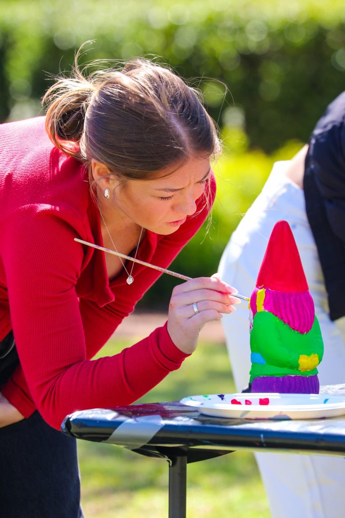 Person painting a brightly coloured garden gnome with a small brush at an outdoor table, with paint palette and greenery in the background.