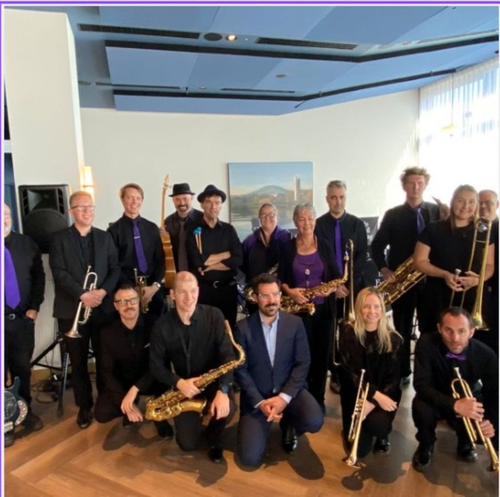 Group of musicians in black outfits posing indoors with brass and woodwind instruments.