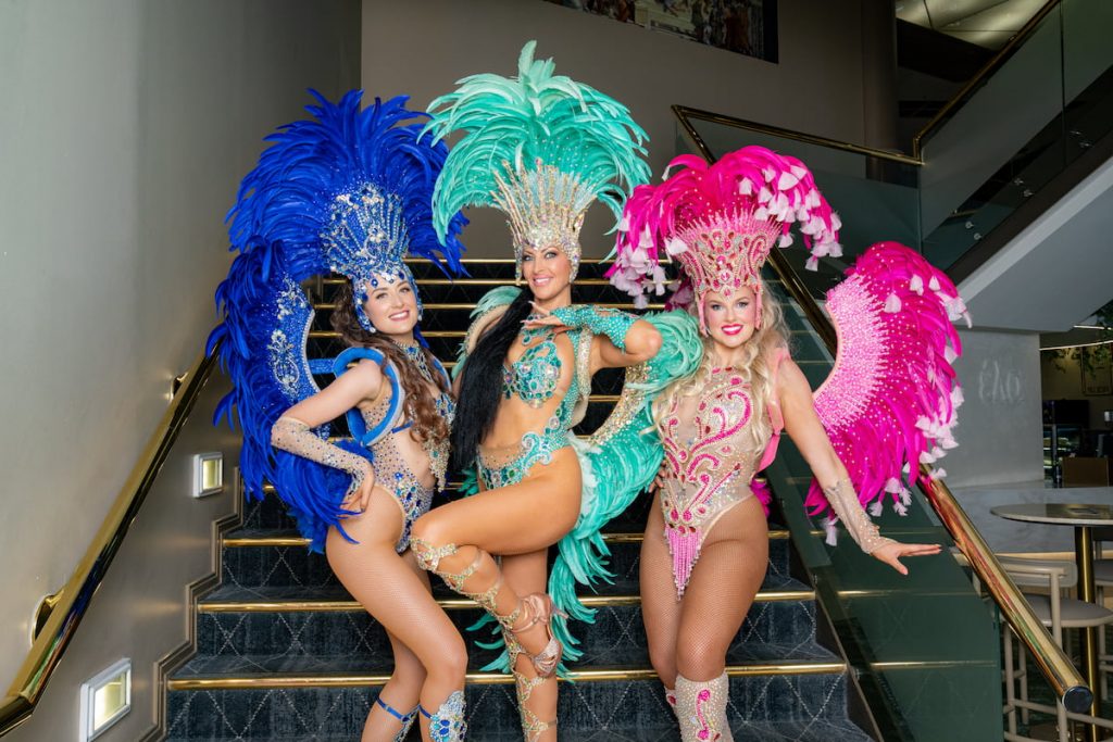 Dancers in bright, feathered costumes perform along a sunny park path with trees and city buildings behind them.