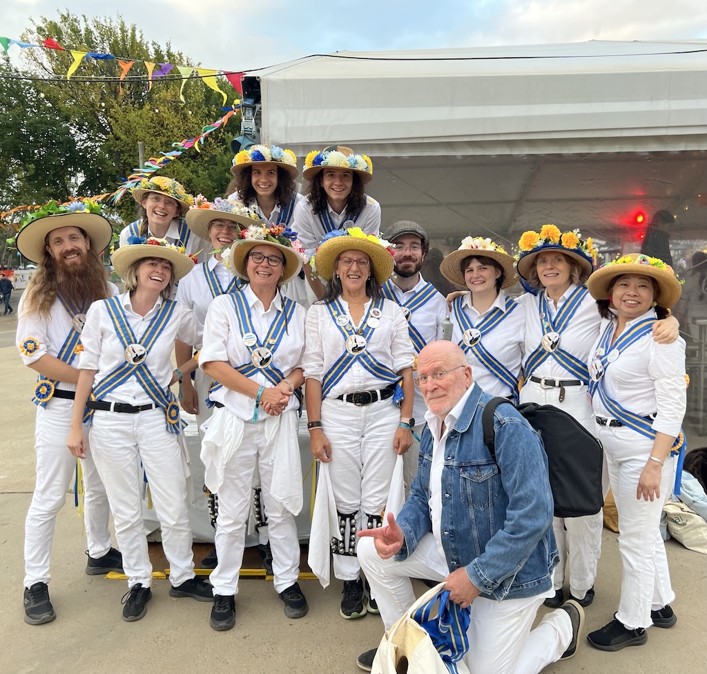 A group in matching white outfits and decorated hats pose together in front of a festival tent.