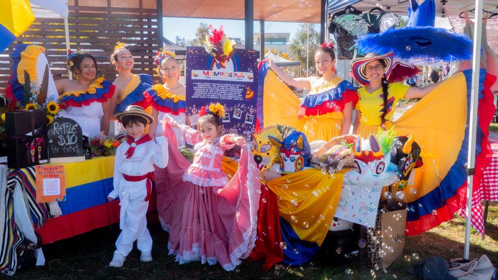 Performers in vibrant costumes pose at a festival stall with decorations, crafts, and flowers.