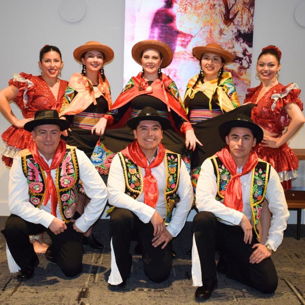 A group in colorful traditional costumes pose indoors, with embroidered outfits and hats.