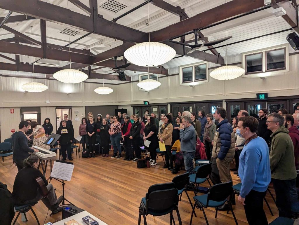 A large group stands in a circle inside a hall with wooden floors and exposed beams, participating in a group singing session while a keyboard player accompanies from the front.
