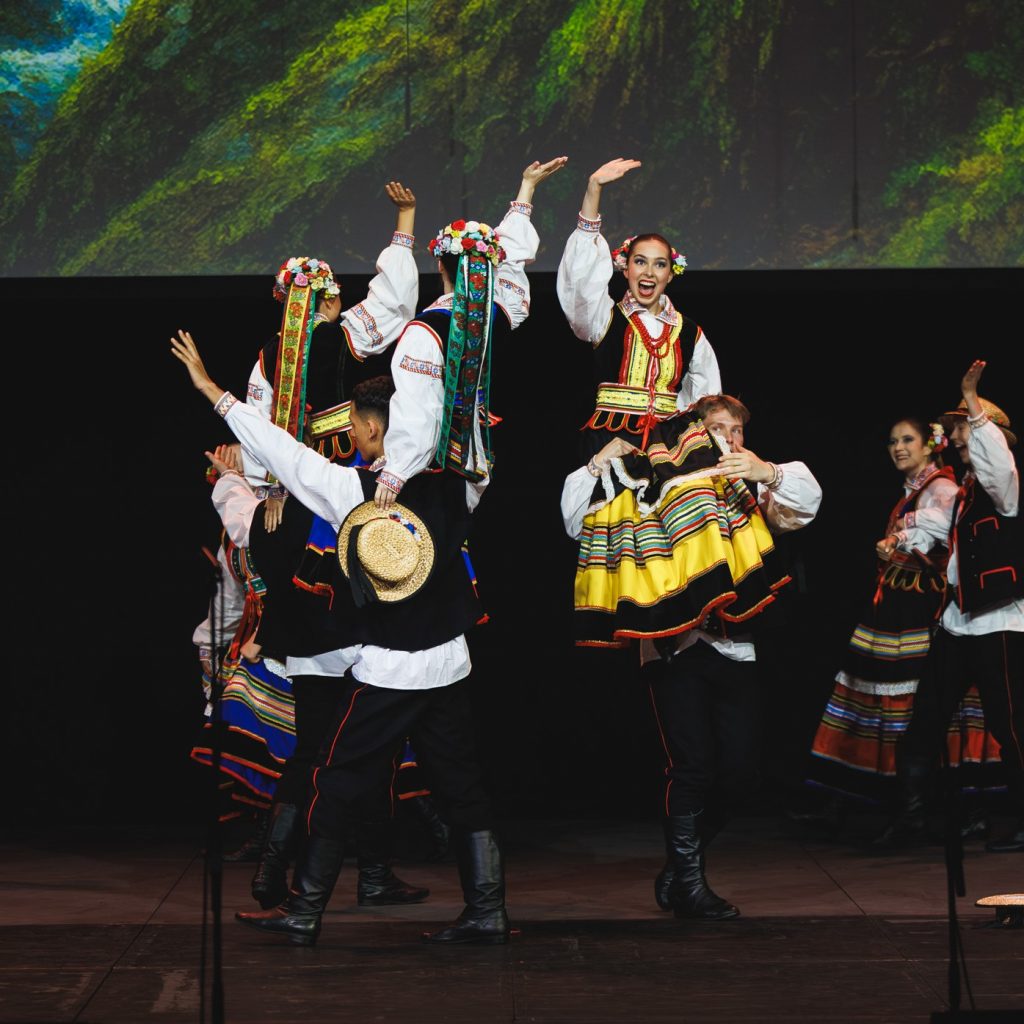 Performers in traditional embroidered costumes dance on stage, with raised arms and patterned skirts against a nature-themed backdrop.