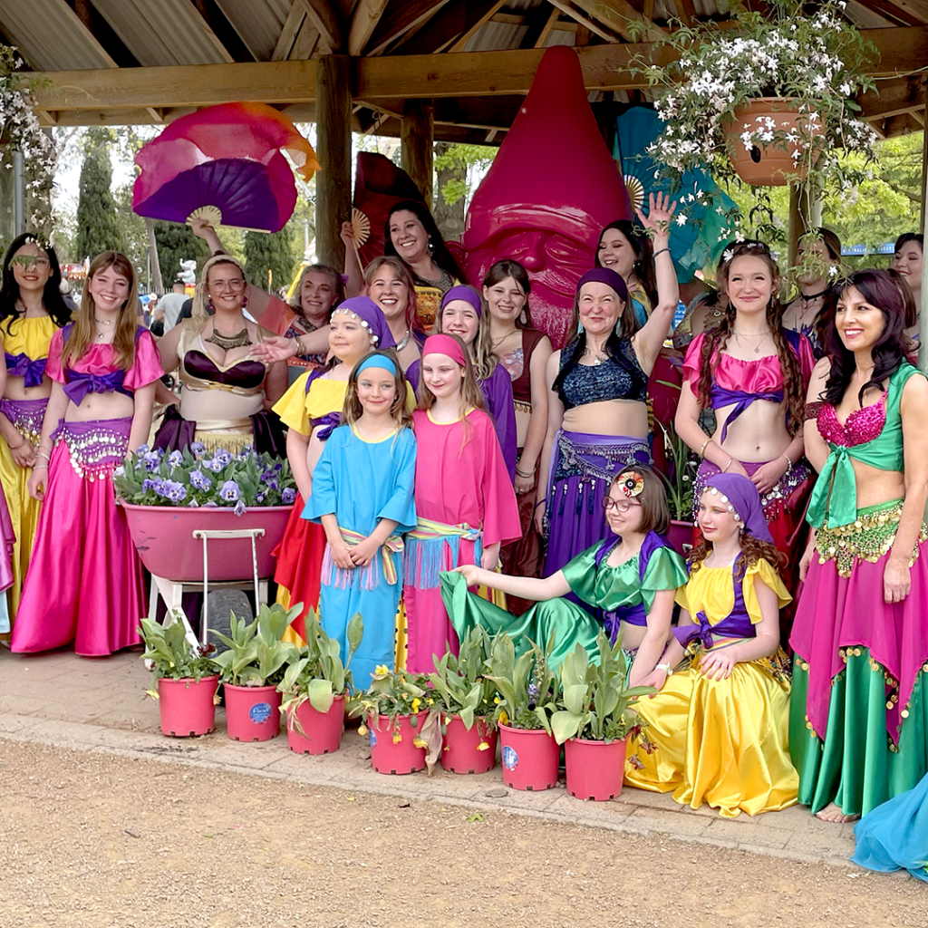 Group in bright, colourful belly dancing costumes posing outdoors around a large pink gnome statue, with potted plants in the foreground.