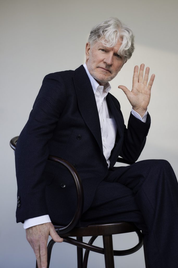 A person with white hair sits on a wooden chair in a dark suit and white shirt, raising one hand in a studio portrait against a plain light background.