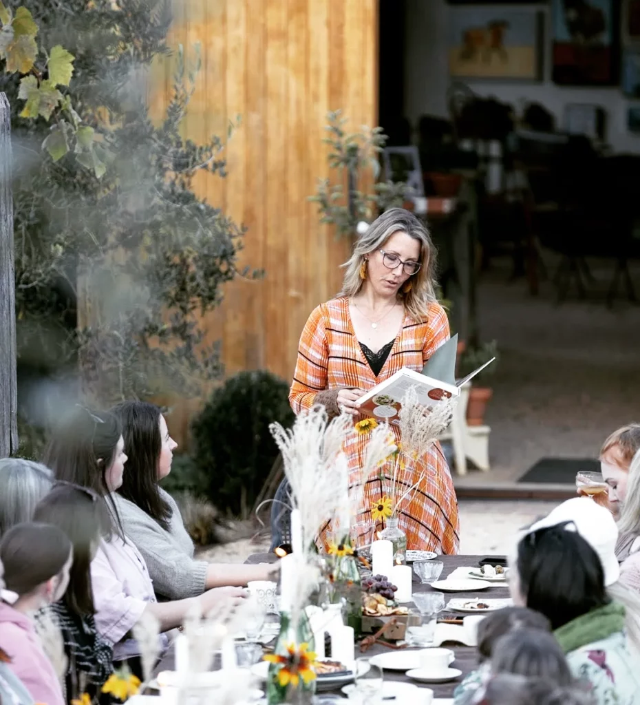 A person in a floral dress stands beside a long outdoor dining table decorated with flowers and candles, while guests gather around in a garden setting.