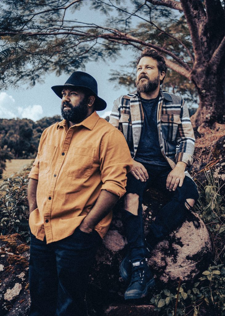 Two musicians stand outdoors beneath trees, looking into the distance in a stylised portrait with warm, muted tones.