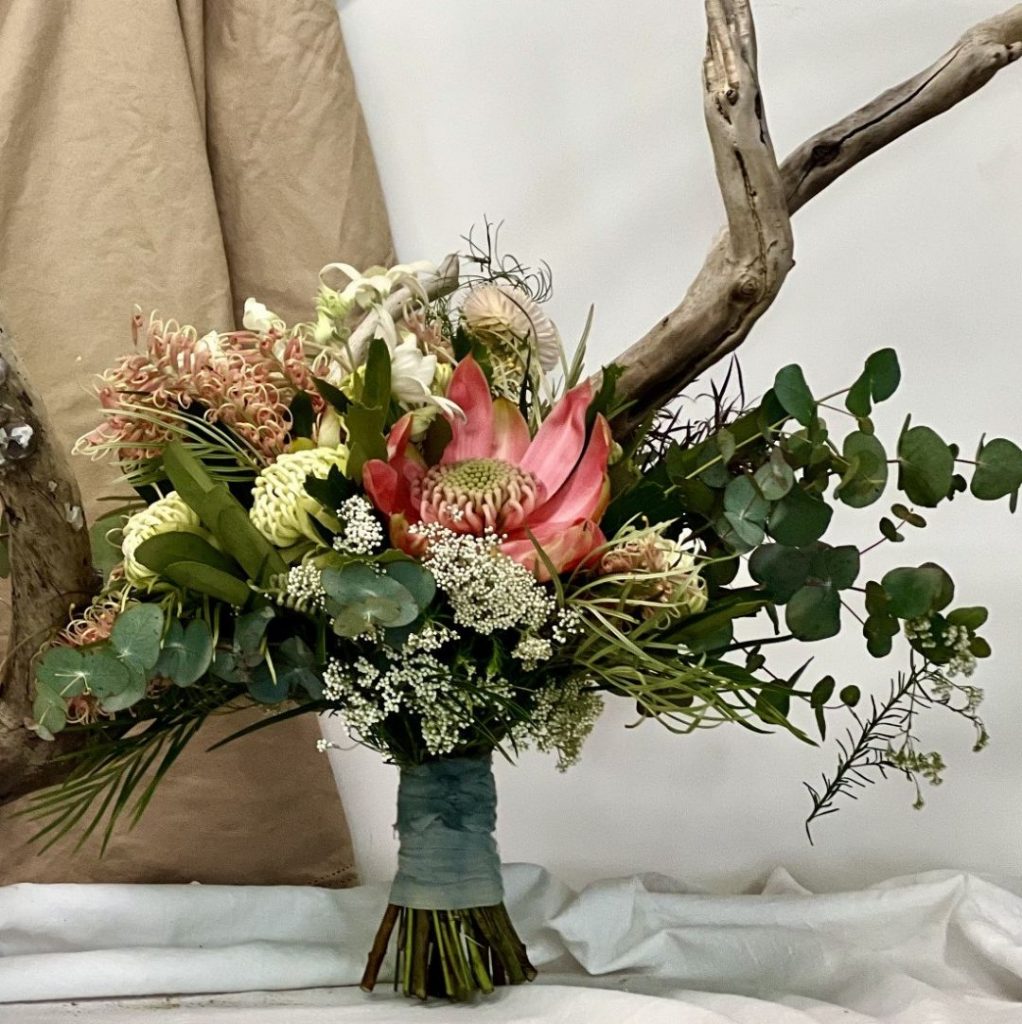 A hand-tied bouquet featuring native flowers and foliage in pink, cream, and green tones, arranged against a neutral backdrop.