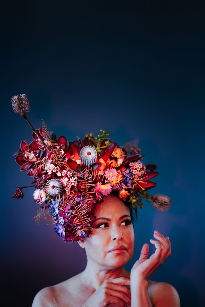 A close-up studio portrait of a performer wearing a dramatic floral headdress of colourful native blooms against a dark background.