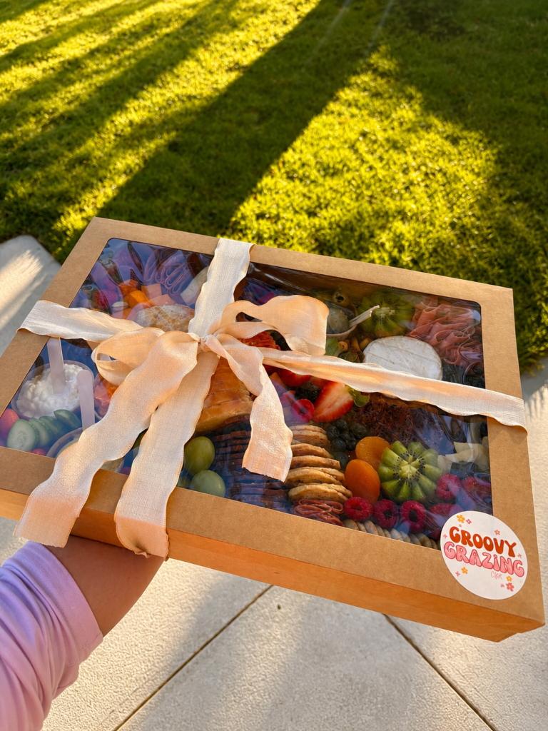 A gourmet grazing box tied with a cream ribbon, held outdoors in warm afternoon sunlight. Through the clear lid, an assortment of cheeses, cured meats, crackers, fresh fruit, dips, and sweet treats is visible. The box is set against a backdrop of green grass and long tree shadows.