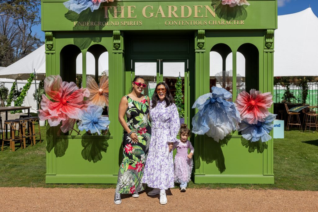 Visitors pose in front of a bright green garden-themed installation decorated with oversized paper flowers at an outdoor festival.