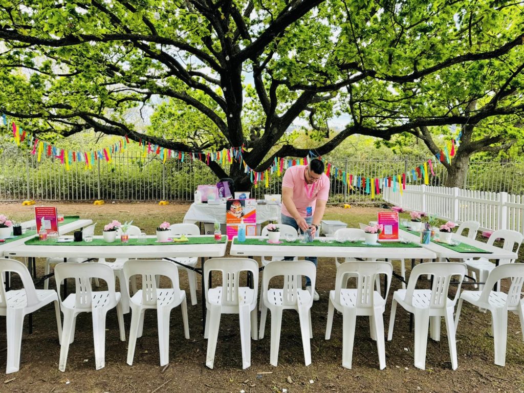 A person bends over long table with white chairs with crafting supplies under a large tree.