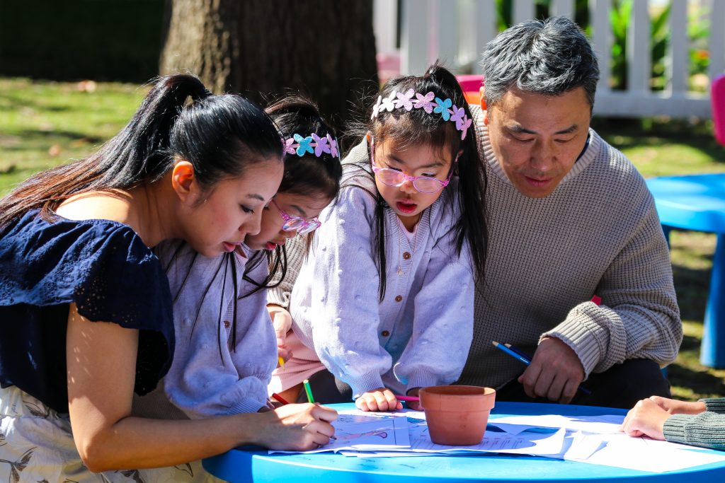 Three children and an older adult gather around a table outdoors, looking closely at an activity, with a tree and sunlight in the background.
