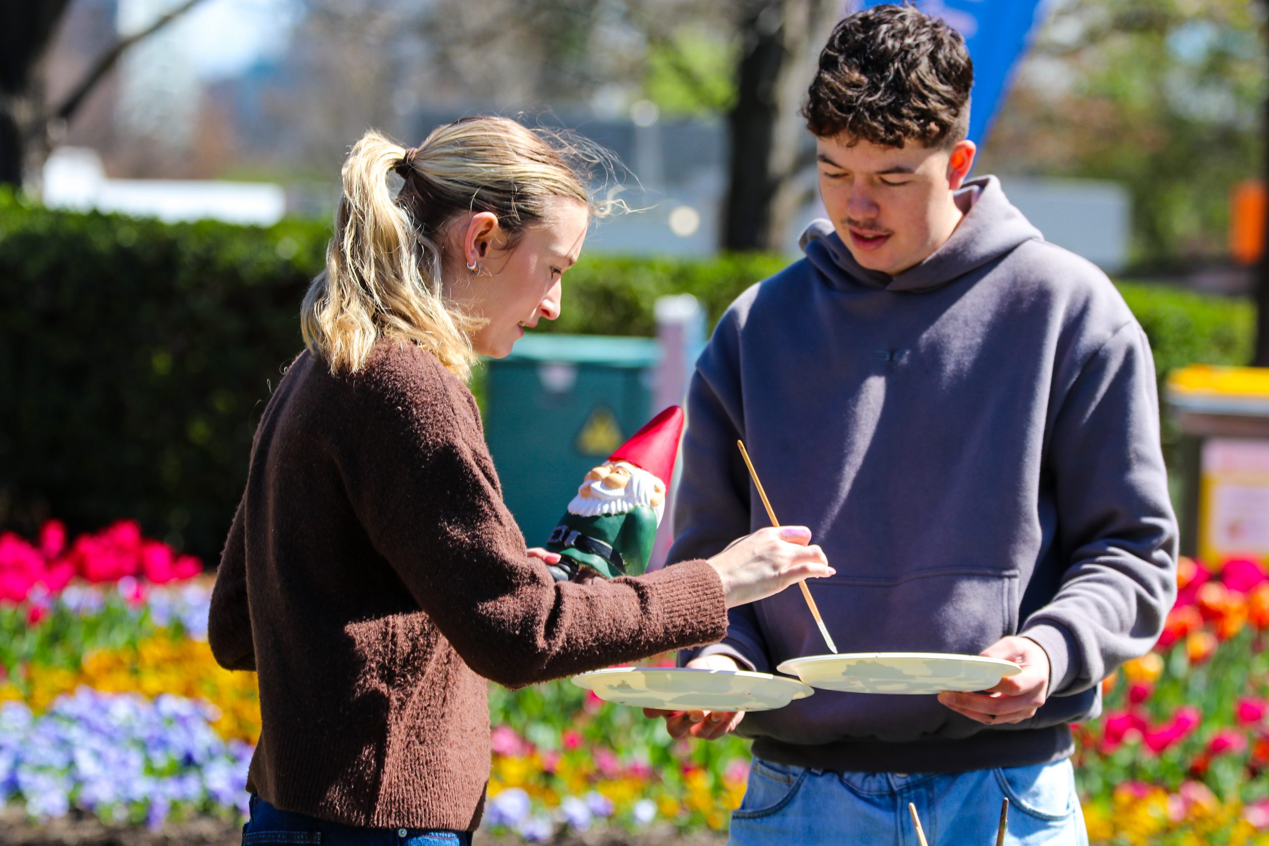 Two people stand beside a flower bed, painting a gnome together outside
