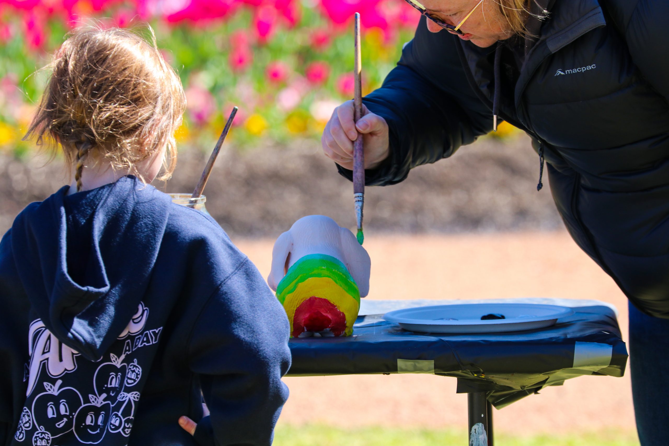 A child paints a brightly coloured rainbow-striped object with a brush at an outdoor craft table.