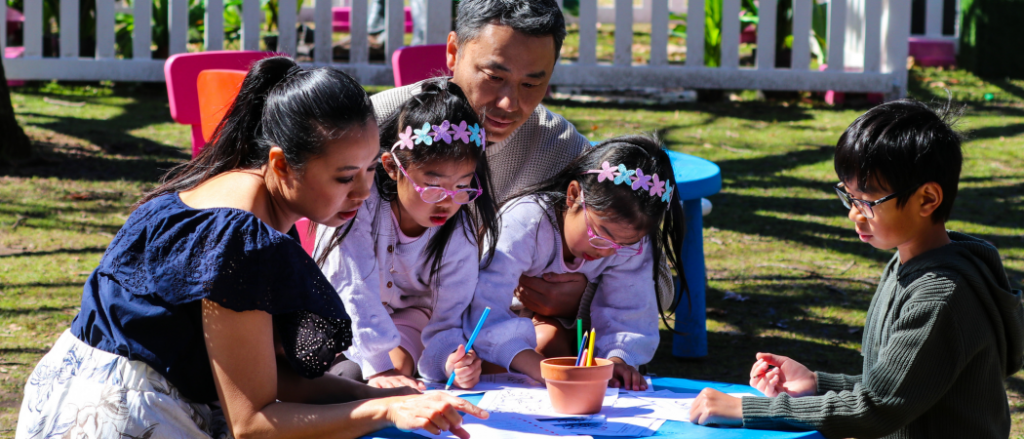 Two adults and three children sit around a small outdoor table, drawing and colouring on activity sheets. The children wear flower headbands and glasses, with coloured pencils and a small terracotta pot on the table in a sunny garden setting.