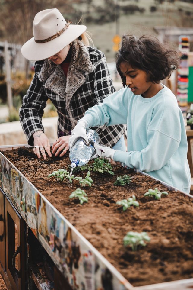 A child and a woman in a hat are planting seeds in a raised garden bed