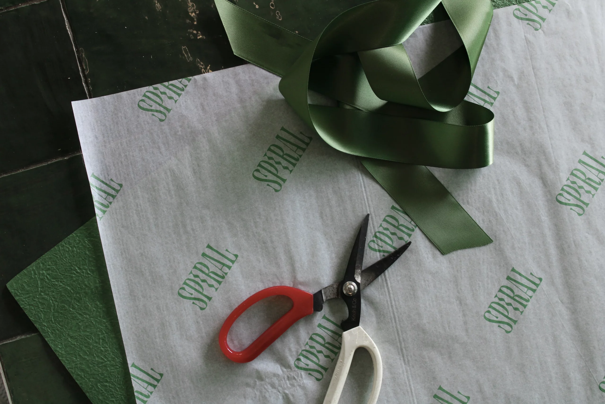 Scissors and ribbon positioned in a dark room over some white and green paper.