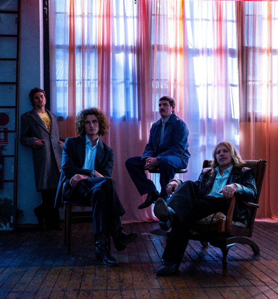 A four-piece band poses for a portrait, featuring the musicians in casual and vintage-inspired clothing against studio backdrop.