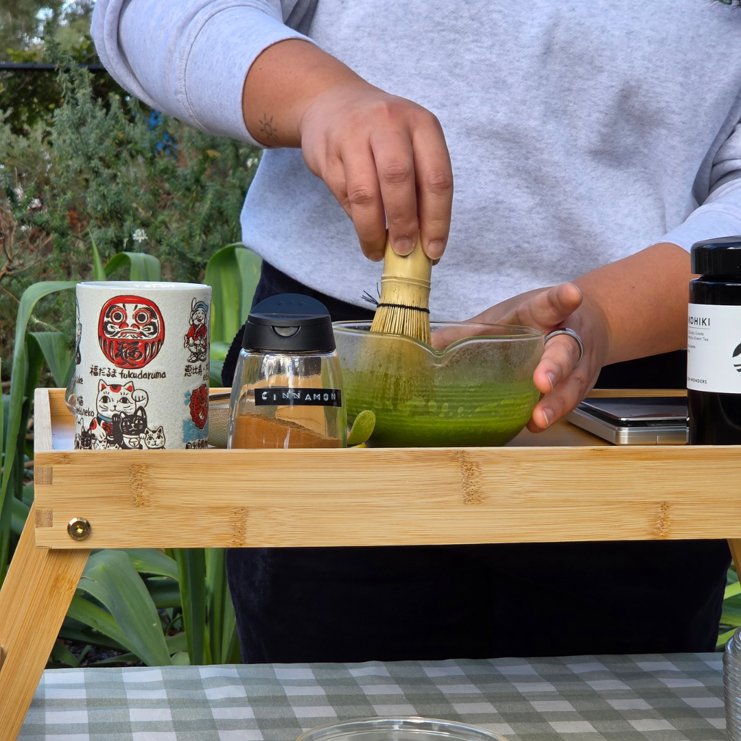 A person whisks vibrant green matcha in a glass bowl on a wooden table, with tea-making accessories and ingredients arranged nearby.