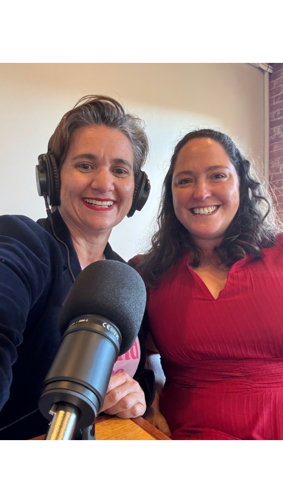 Portrait-oriented image of two people seated side by side at a table with a professional podcast microphone in the foreground. One person is wearing studio headphones, and both are positioned in front of a plain wall with exposed brick visible to one side.