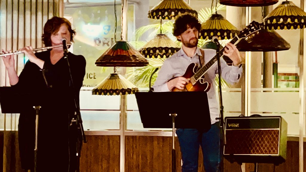 Two musicians perform live indoors, with one playing a flute and the other a guitar, surrounded by decorative hanging lamps in a warmly lit venue.