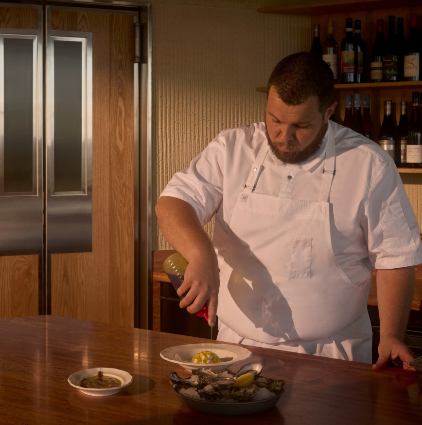 Head Chef Will Moyle, dressed in a white chef's jacket and apron, plates a dish at a wooden counter. Oysters, a lemon wedge and small dishes are arranged in the foreground, while wine bottles line shelves in the background.