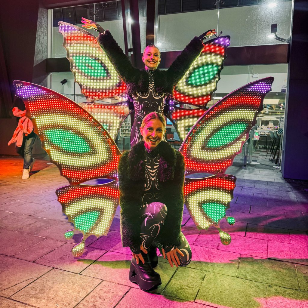 Promotional image of two performers wearing large LED butterfly wings displaying red, green, yellow, and purple light patterns. One performer stands with raised arms while the other kneels in front.