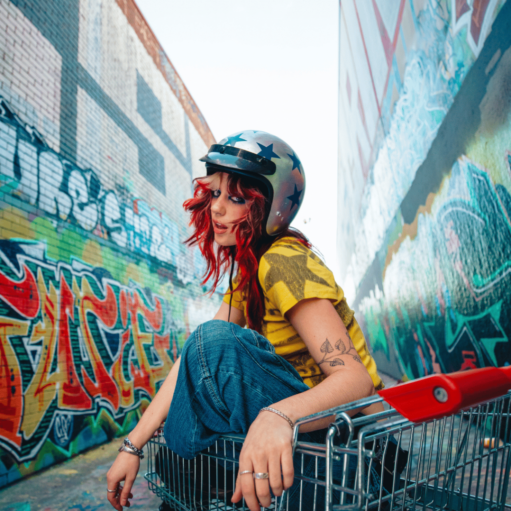 Promotional image of a person seated in a shopping trolley between two walls covered in colourful street art. The person wears a black helmet, a yellow shirt, and wide-leg blue jeans.