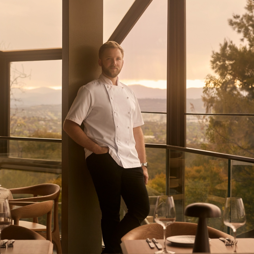 Lunetta Executive Chef Tristan Rebbettes stands beside floor-to-ceiling windows in a restaurant dining room, with set tables in the foreground and scenic views of trees and distant hills beyond.